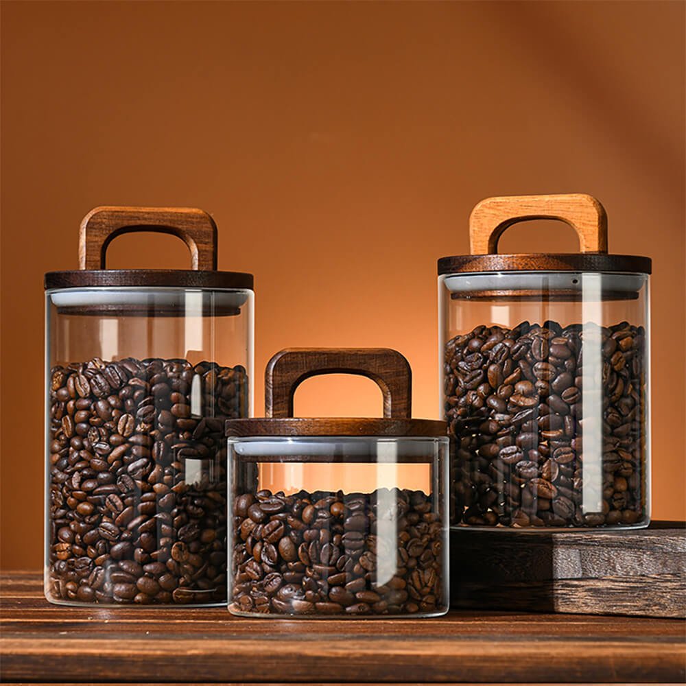 A set of transparent pantry jar storage containers filled with coffee beans, featuring cylindrical glass bodies and dark wooden lids with built-in handles. The jars are arranged on a wooden surface against a warm-toned background, varying in size to create a visually appealing display.