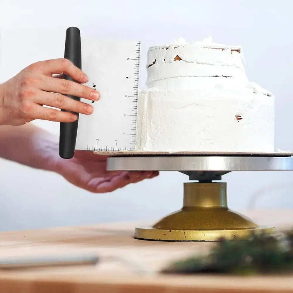 A hand uses a pastry scraper to smooth icing on a cake set on a rotating stand in a kitchen workspace.