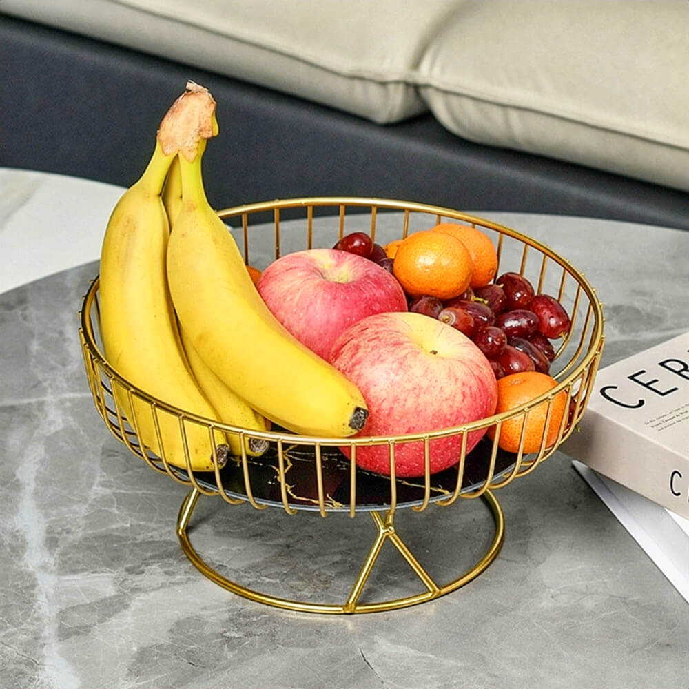 A pedestal fruit bowl with a black marble base and gold metal wire frame, elegantly displaying a variety of fresh fruits including bananas, apples, grapes, and oranges. The bowl is placed on a gray marble table, adding a sophisticated touch to the setting.