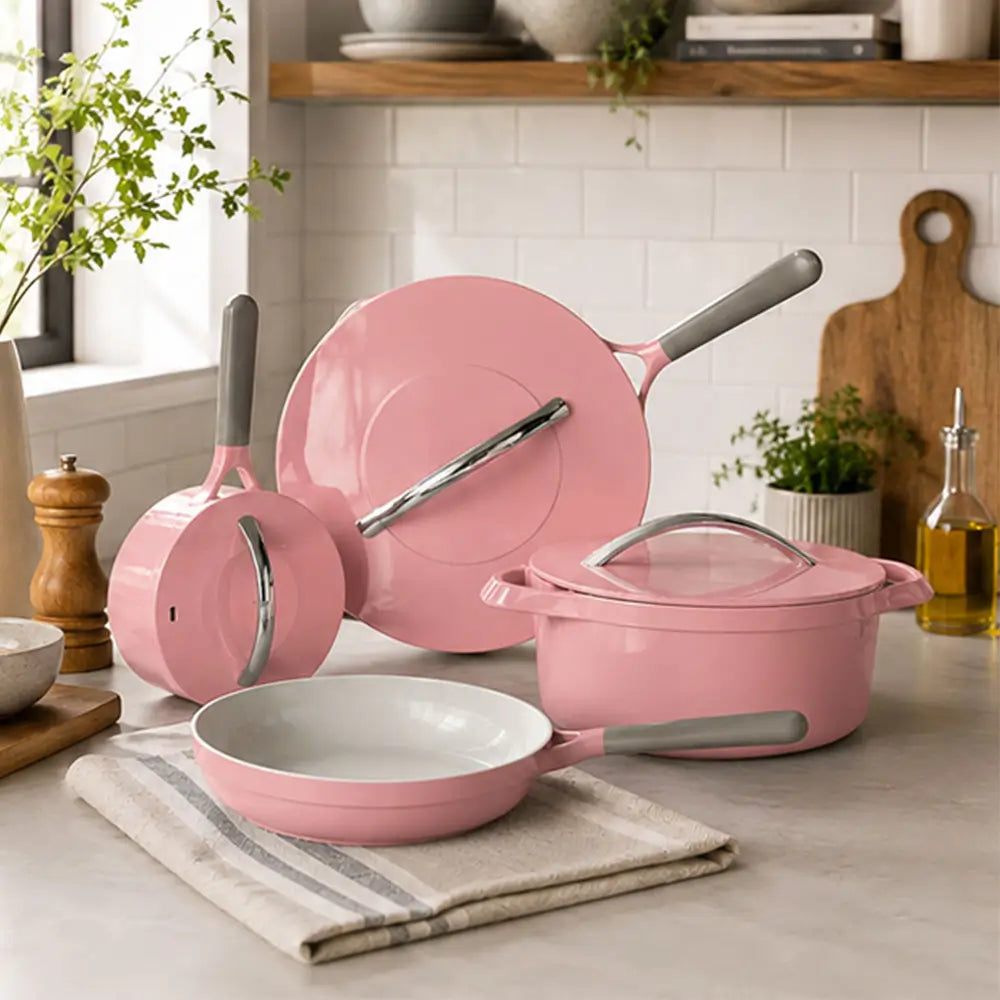 Pink ceramic cookware set including a frying pan, saucepan, stockpot, and glass lid arranged on a kitchen countertop.
