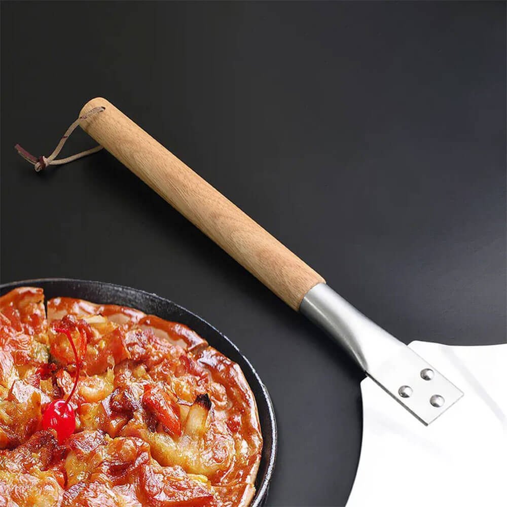 Close-up of a pizza peel with a detachable oak handle, placed next to a delicious deep-dish pizza on a black surface.