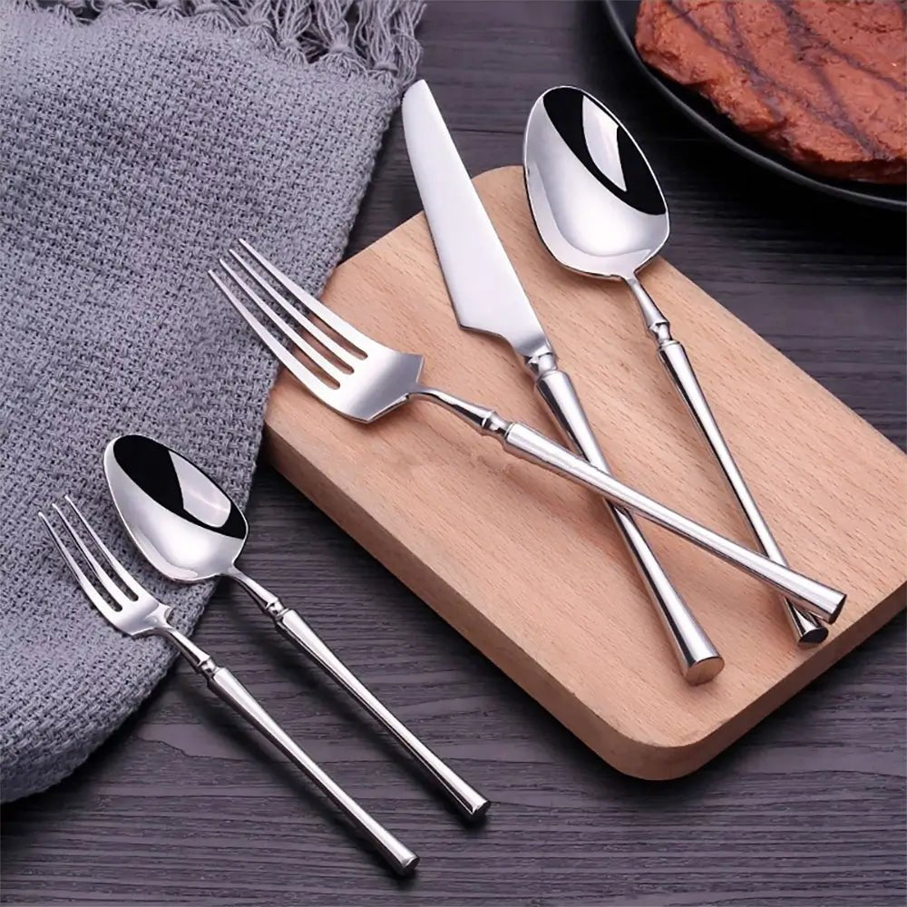 Stainless steel silver set with knife, forks, and spoons on black wood and slate with white flowers and a teacup nearby.