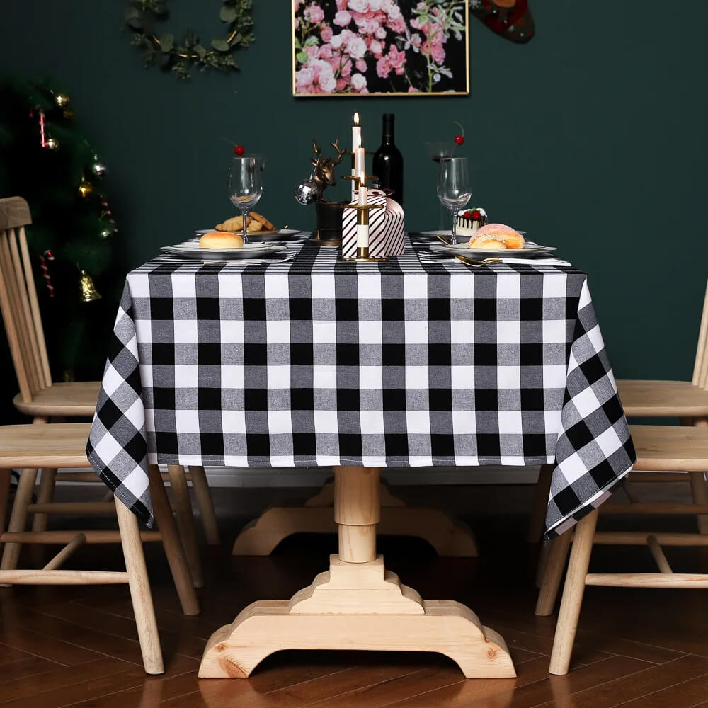 A dining table is styled with a classic black and white plaid Christmas fabric tablecloth, perfect for festive gatherings. The table is set with elegant wine glasses, white plates, and gold utensils, complemented by small desserts and a decorative centerpiece featuring candles and a reindeer figurine. The green wall background, floral artwork, and Christmas tree add to the warm and celebratory ambiance.