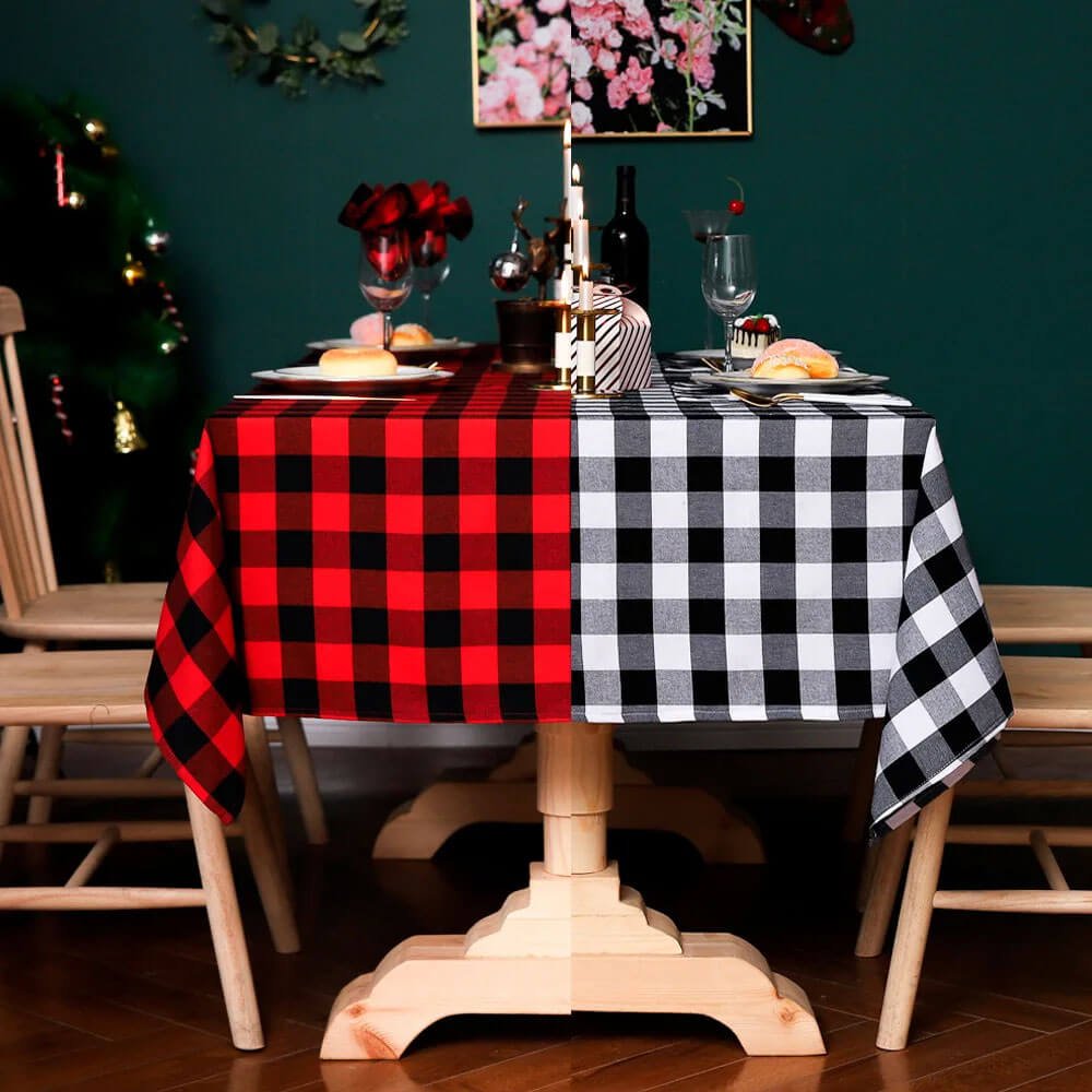 A dining table is decorated with a red and black plaid tablecloth on one side and a black and white plaid tablecloth on the other. The table is set with plates, glasses, and festive decorations. The setup is complemented by a wooden table and chairs, with a dark green background and a Christmas tree adding to the festive vibe.