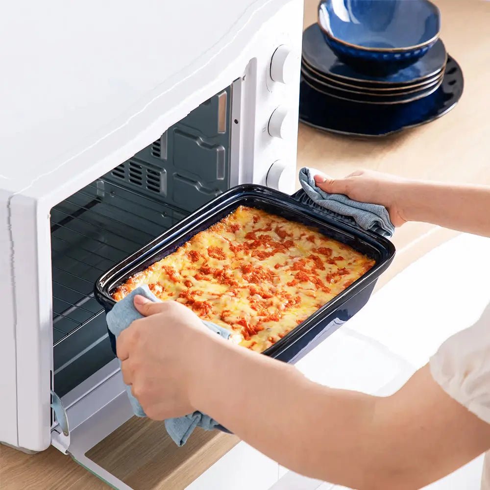 Blue porcelain casserole dish filled with baked lasagna being taken out of an oven using cloth holders.