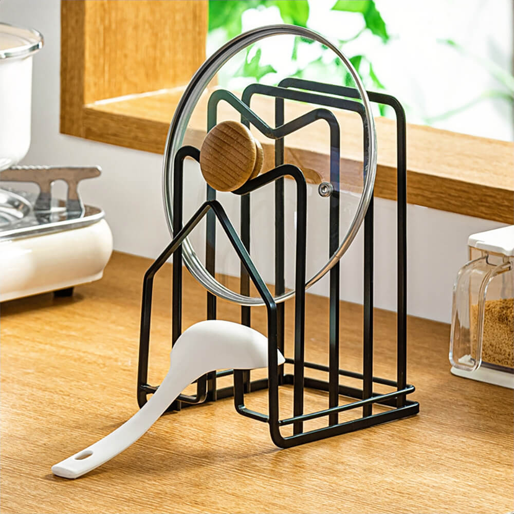 A versatile black rack for cutting boards, featuring a modern house-shaped metal design. It efficiently holds multiple cutting boards upright and includes space to accommodate a pot lid and a cooking utensil, as shown with a glass lid and a white spoon. Placed on a wooden kitchen countertop with natural light, it enhances both functionality and aesthetics in the kitchen.