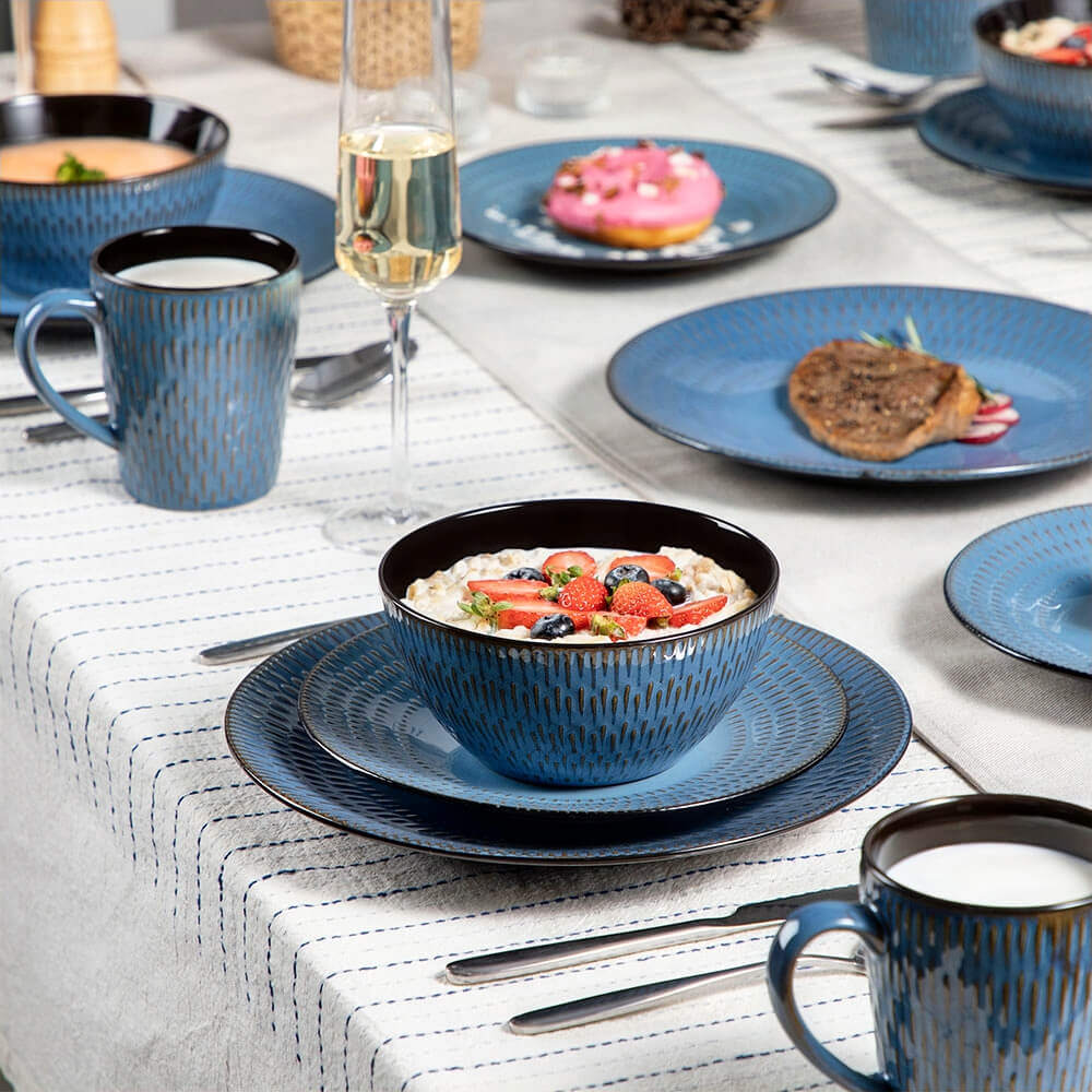 A dining table set with reactive glaze stoneware dinnerware sets in a rich blue finish. The table features matching plates, bowls, and mugs with embossed designs and sleek black interiors. A bowl filled with oatmeal, strawberries, and blueberries is set alongside other plates with various dishes, complemented by utensils and a champagne glass on a striped tablecloth.