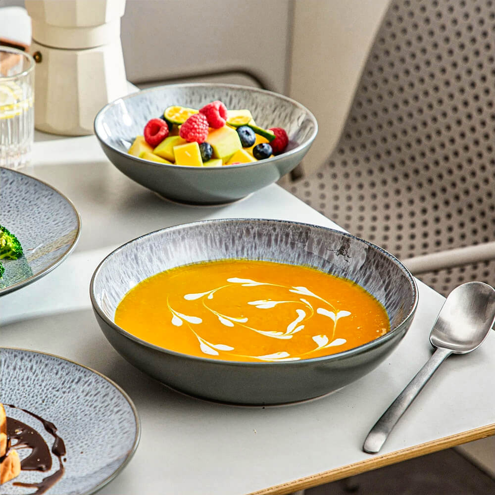 A close-up of a reactive glaze stoneware dinnerware set, showcasing its speckled gray-blue design. The bowl elegantly holds a vibrant orange soup with artistic cream swirls, complemented by a matching fruit bowl filled with mango, berries, and kiwi. A sleek silver spoon rests beside the set, enhancing the minimalist yet sophisticated dining presentation.