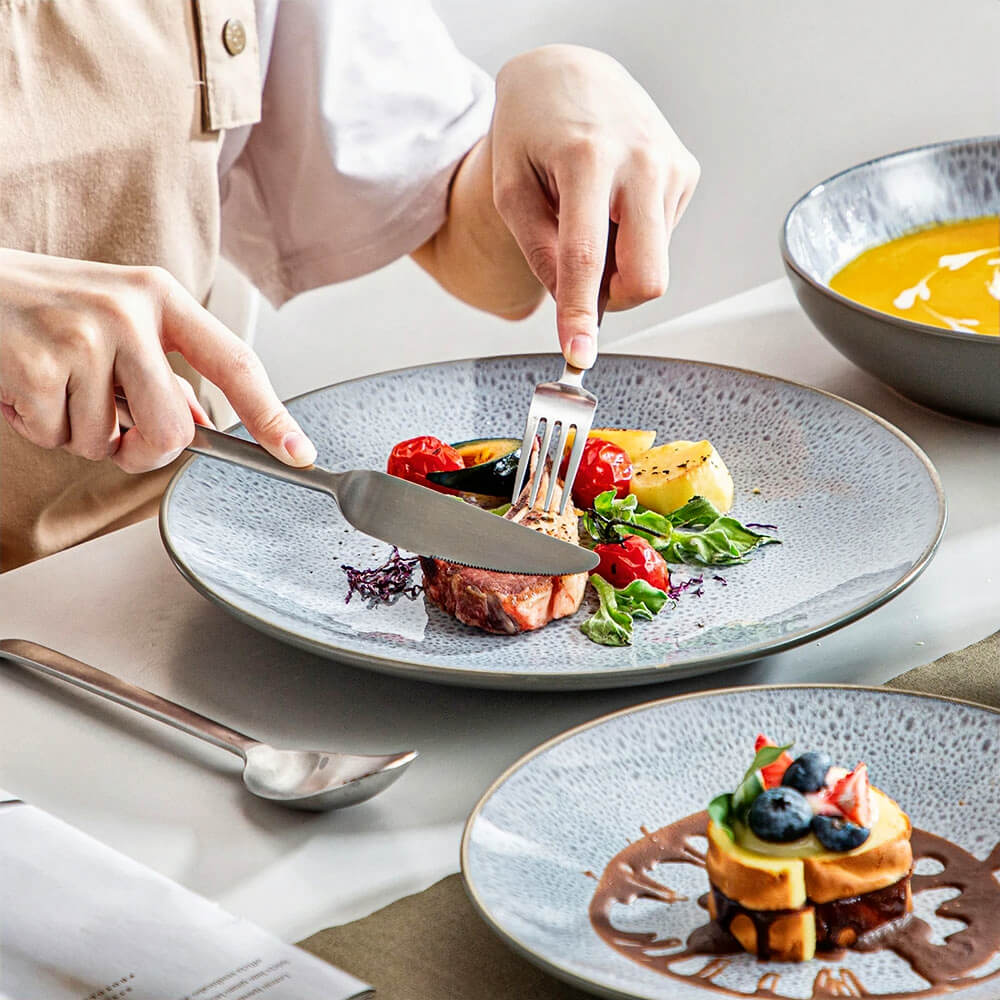 A person dining with a reactive glaze stoneware plate, its speckled gray-blue finish adding a touch of elegance. The plate features a beautifully arranged meal of roasted vegetables, fresh greens, and a tender cut of meat. A matching bowl of vibrant orange soup is visible in the background, complementing the set.