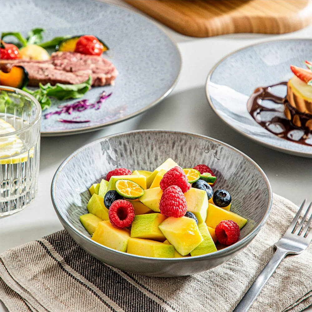 A vibrant fruit salad served in a reactive glazed stoneware bowl, featuring a speckled gray-blue finish that adds an artistic touch. The salad includes fresh mango, raspberries, blueberries, and slices of kumquat, creating a colorful and appetizing presentation. The bowl is set on a striped napkin, surrounded by matching stoneware plates showcasing elegantly plated meals, completing the stylish and modern table setting.