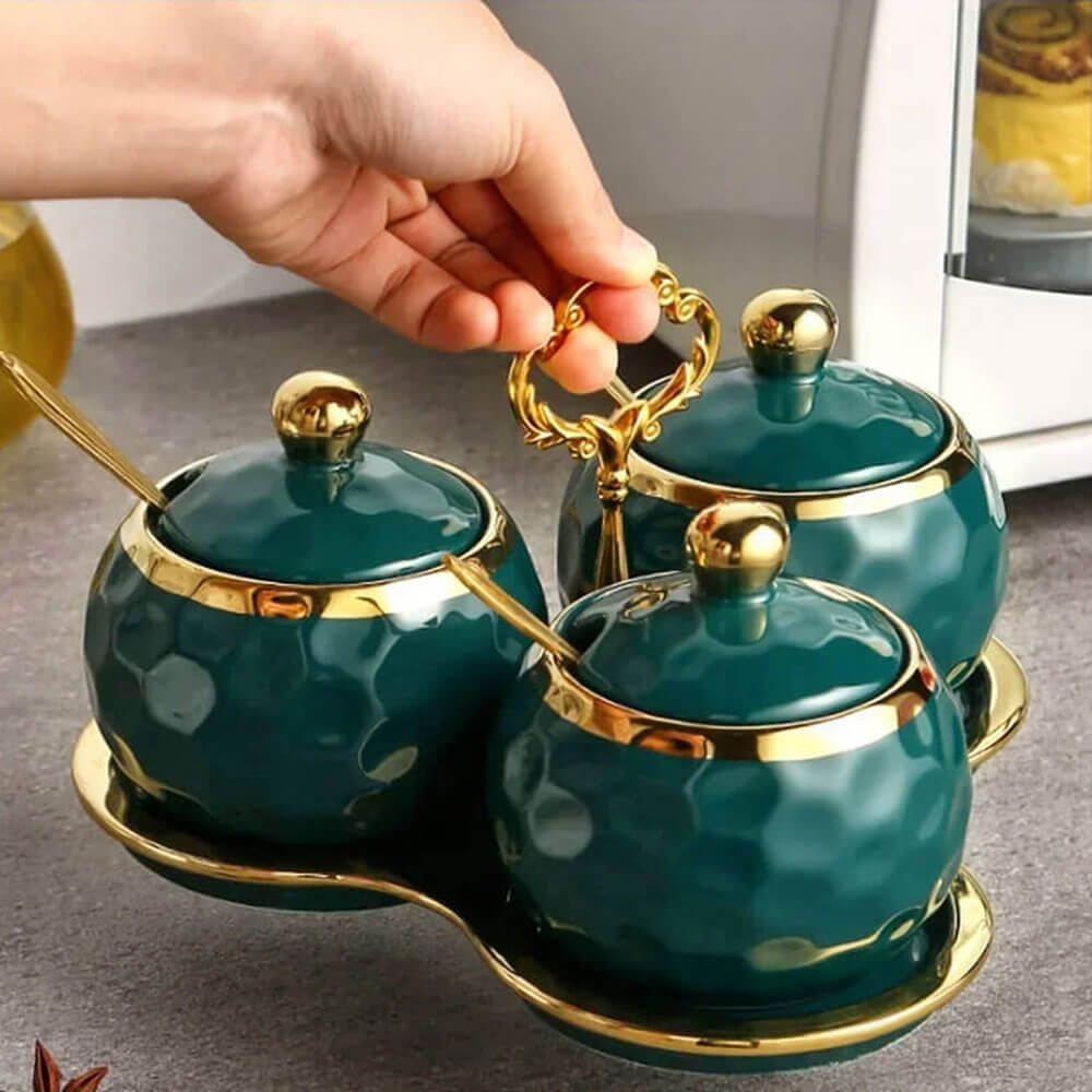 A hand lifting a set of reusable condiment containers with gold-trimmed lids, matching spoons, and a decorative gold-handled tray.