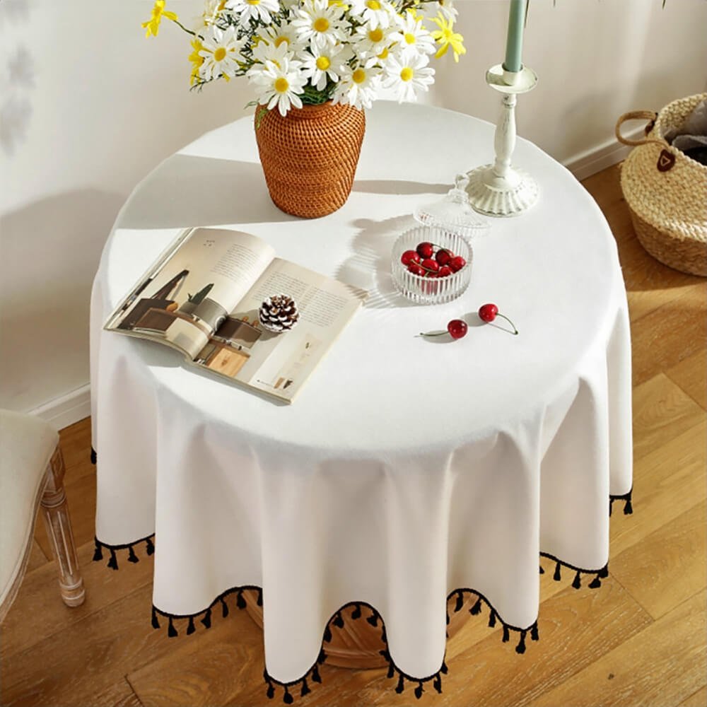 A round tablecloth with tassels in white elegantly drapes over a small round table. The tablecloth features black tassels along the edges, adding a decorative and stylish touch. The table is adorned with a woven vase filled with white and yellow daisies, a white candle holder with a candle, a crystal dish with cherries, and an open book with a pinecone resting on it.