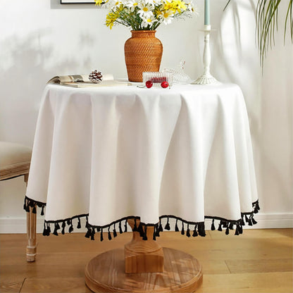 A round tassel tablecloth in white covers a small round wooden table. The tablecloth features black tassels along the edge for a decorative accent. The table is styled with a woven vase holding white and yellow daisies, a white candle holder with a candle, a pinecone, an open book, and small decorative items, creating a cozy and elegant decor setup.