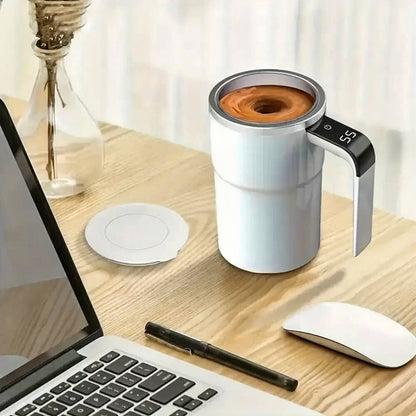 A self-stirring coffee mug is placed on a wooden desk beside a laptop, pen, and wireless mouse. The mug is actively stirring the beverage, creating a vortex, with the temperature displayed on the LED screen embedded in the handle. The lid is set aside, showcasing the mug&