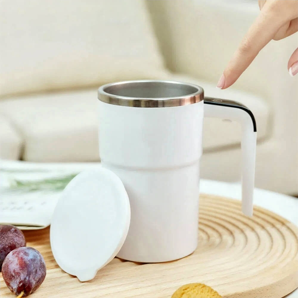 A white self-stirring mug with a sleek stainless steel rim is displayed on a wooden surface. The lid is placed beside the mug, and a person is activating the stirring function by pressing the touch button on the handle.