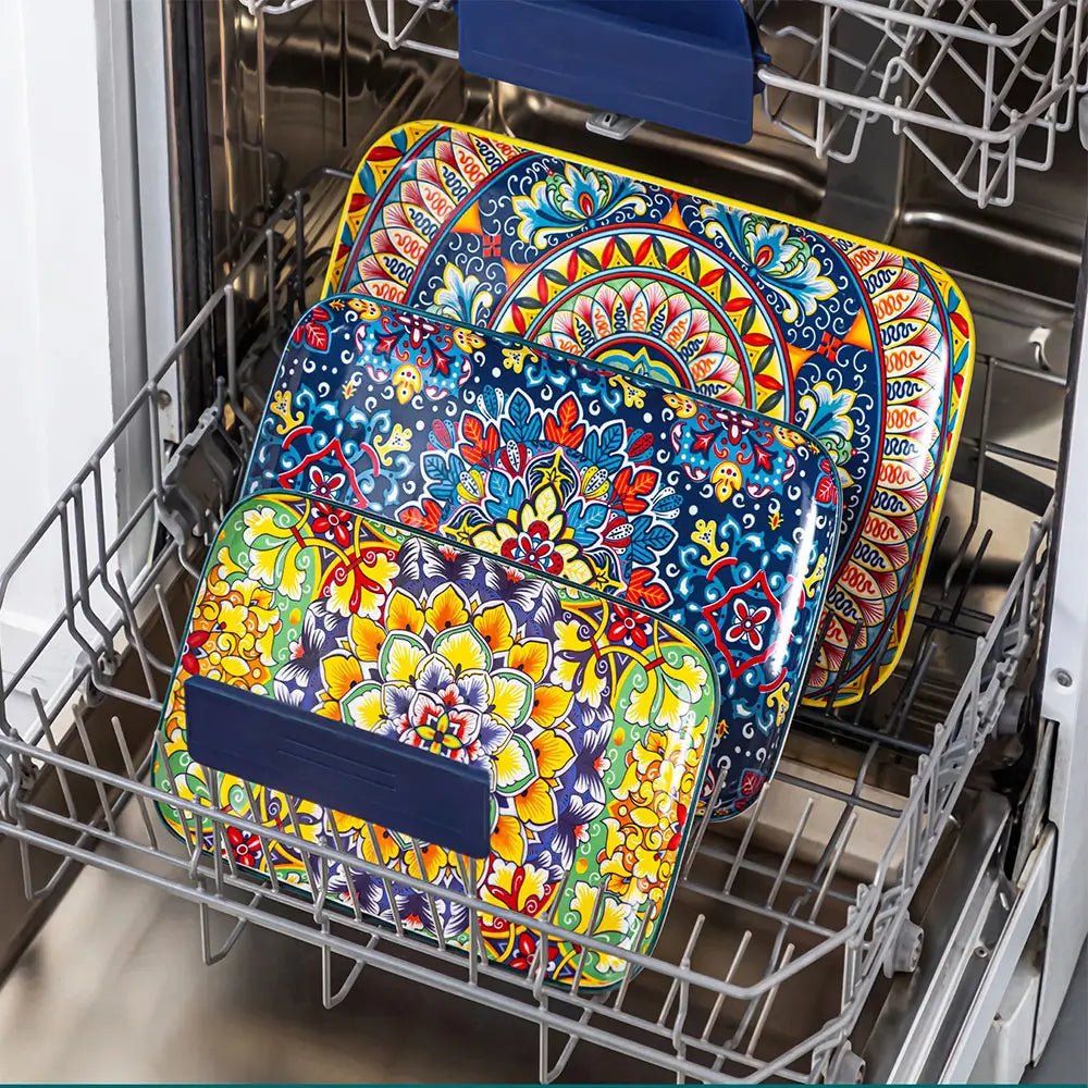 Colorful ceramic serving plates with floral and geometric patterns placed inside a dishwasher rack for cleaning.
