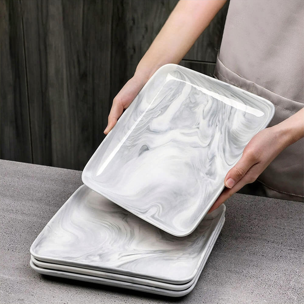 A person wearing a beige apron holds a marble-style serving plate from a stacked set on a gray countertop. The plates have a square shape, rounded edges, and a glossy gray and white swirl pattern.