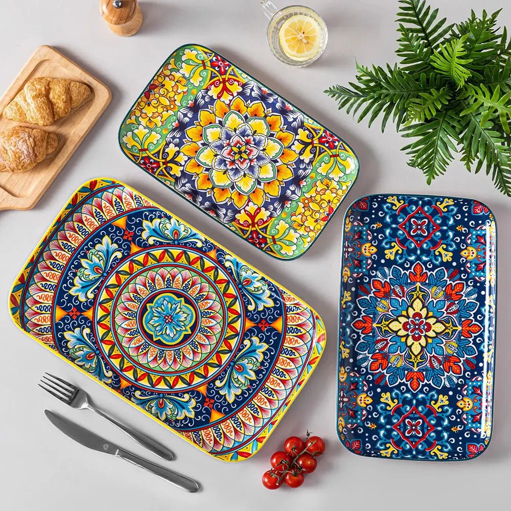 Three rectangular serving plates with colorful floral and geometric patterns placed on a dining table with food, cutlery, and a drink.
