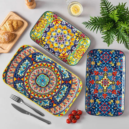 Three rectangular serving plates with colorful floral and geometric patterns placed on a dining table with food, cutlery, and a drink.