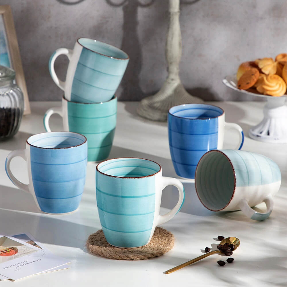 A set of six stoneware mugs in varying shades of blue and green arranged on a white table. Each mug features a subtle striped design and a smooth finish, with a slight rustic touch along the rim. Some mugs are standing upright, while others are stacked or lying on their sides. The scene is including a plate of cookies, a decorative lamp, coffee beans, and a golden spoon.