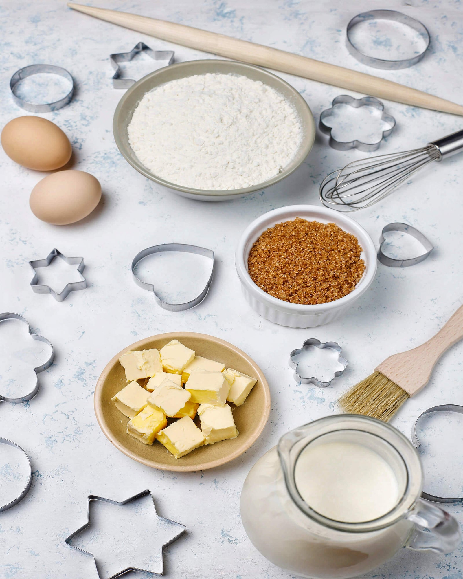 A baking setup featuring ingredients and tools, including flour, brown sugar, butter, eggs, and milk, arranged with cookie cutters, a whisk, a rolling pin, and a pastry brush on a textured surface. Perfect for a shop bakeware promotion.