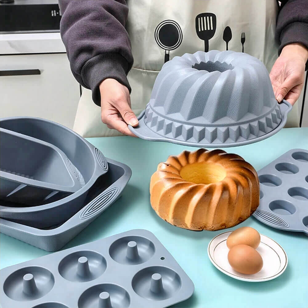 Person holding a gray silicone bundt pan surrounded by various silicone baking molds, baked cake, and eggs on a kitchen counter.