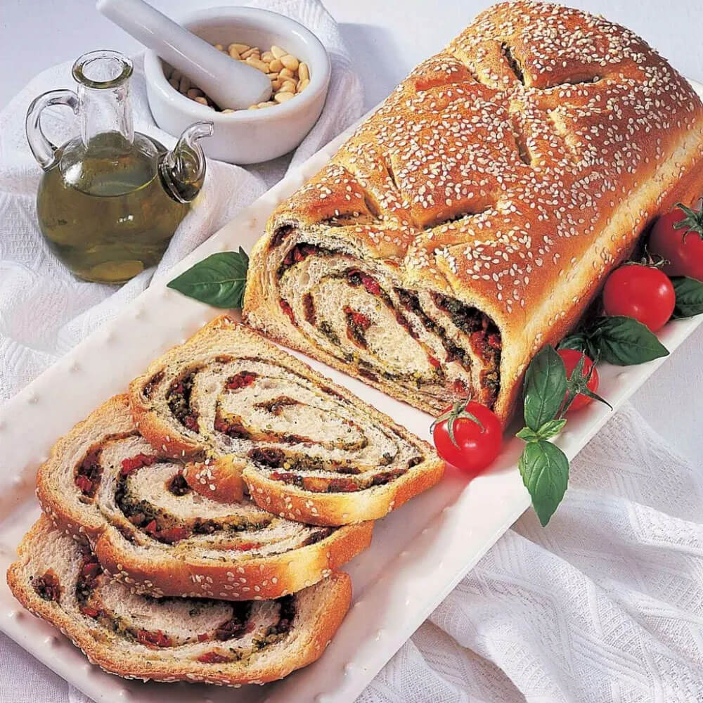 A freshly baked loaf of bread on a white rectangular serving plate. The bread is sliced to reveal a swirl of savory filling, featuring herbs, sun-dried tomatoes, and pesto. The loaf is topped with sesame seeds and garnished with fresh basil leaves and cherry tomatoes. A bottle of olive oil and a mortar with pine nuts are in the background.