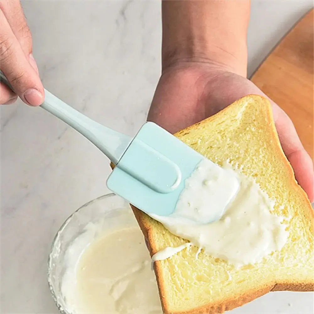 Person spreading cream onto a slice of toast using a blue silicone spatula above a bowl of creamy mixture.