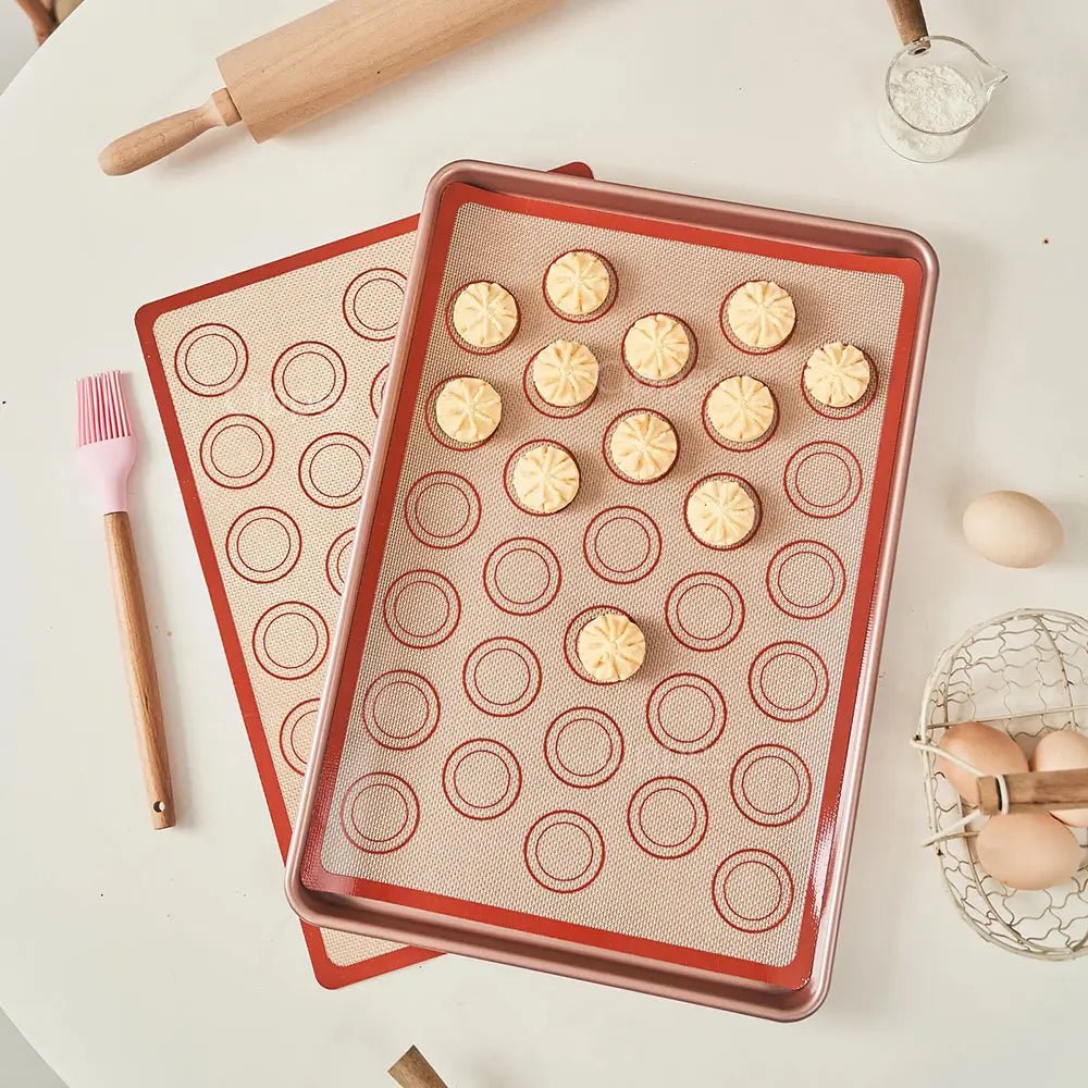 Silicone baking mats with macaron shells on a white surface with baking tools.