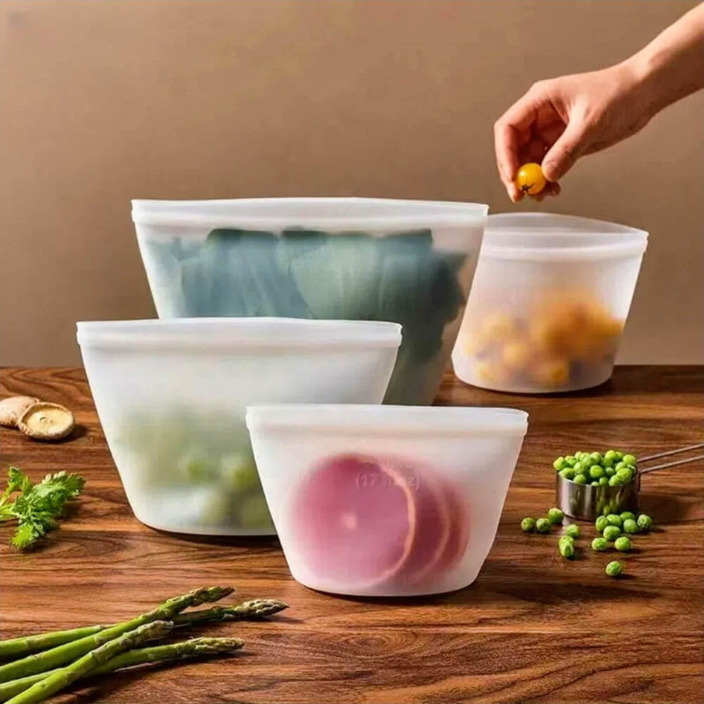Four translucent silicone reusable bags filled with fresh produce including leafy greens, tomatoes, peas, and lemon slices