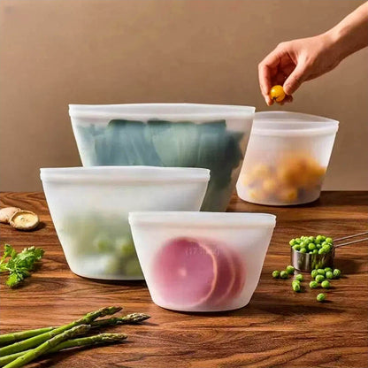 Four translucent silicone reusable bags filled with fresh produce including leafy greens, tomatoes, peas, and lemon slices
