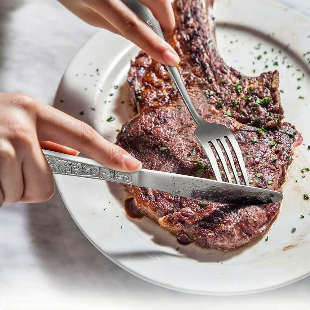 Silver flatware set in use, featuring an intricately designed knife and fork cutting into a perfectly cooked steak on a white plate.