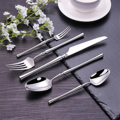 Silverware set with knife, forks, and spoons on wood board and gray cloth, served beside grilled meat on black plate.