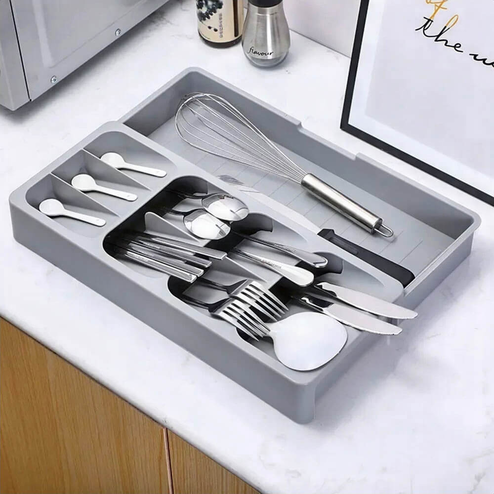 A gray silverware drawer organizer with an expandable design, neatly storing spoons, forks, knives, and utensils. Placed on a white countertop alongside a whisk, framed wall art, and salt and pepper shakers for a well-organized kitchen setup.