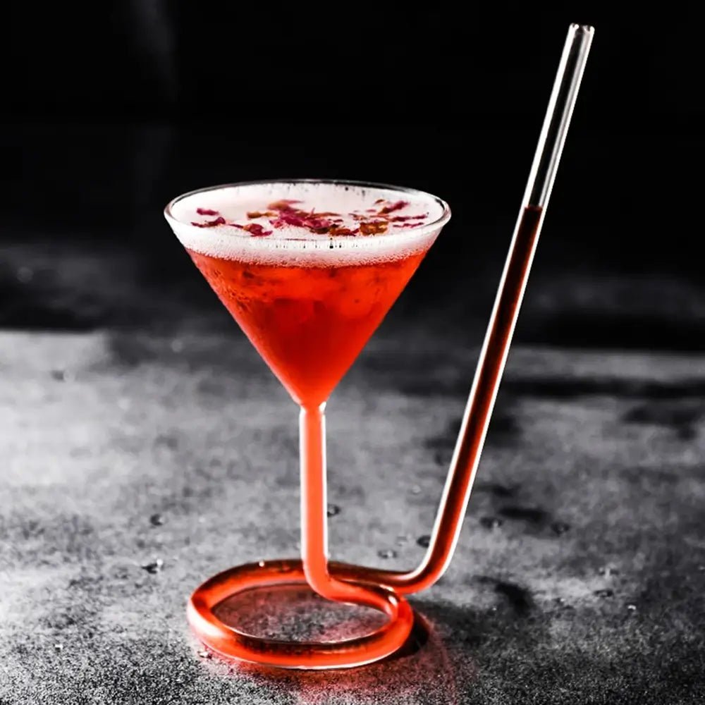 Spiral cocktail glass with built-in straw stem, holding a red drink topped with foam and petals, placed on a dark surface.