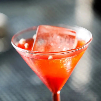 Red cocktail in a martini glass with a large ice cube on a dark surface