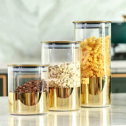 Three airtight stackable glass canisters with gold accents are placed on a kitchen countertop. The canisters are filled with coffee beans, oats, and spiral pasta, highlighting their elegant and practical design for food storage.