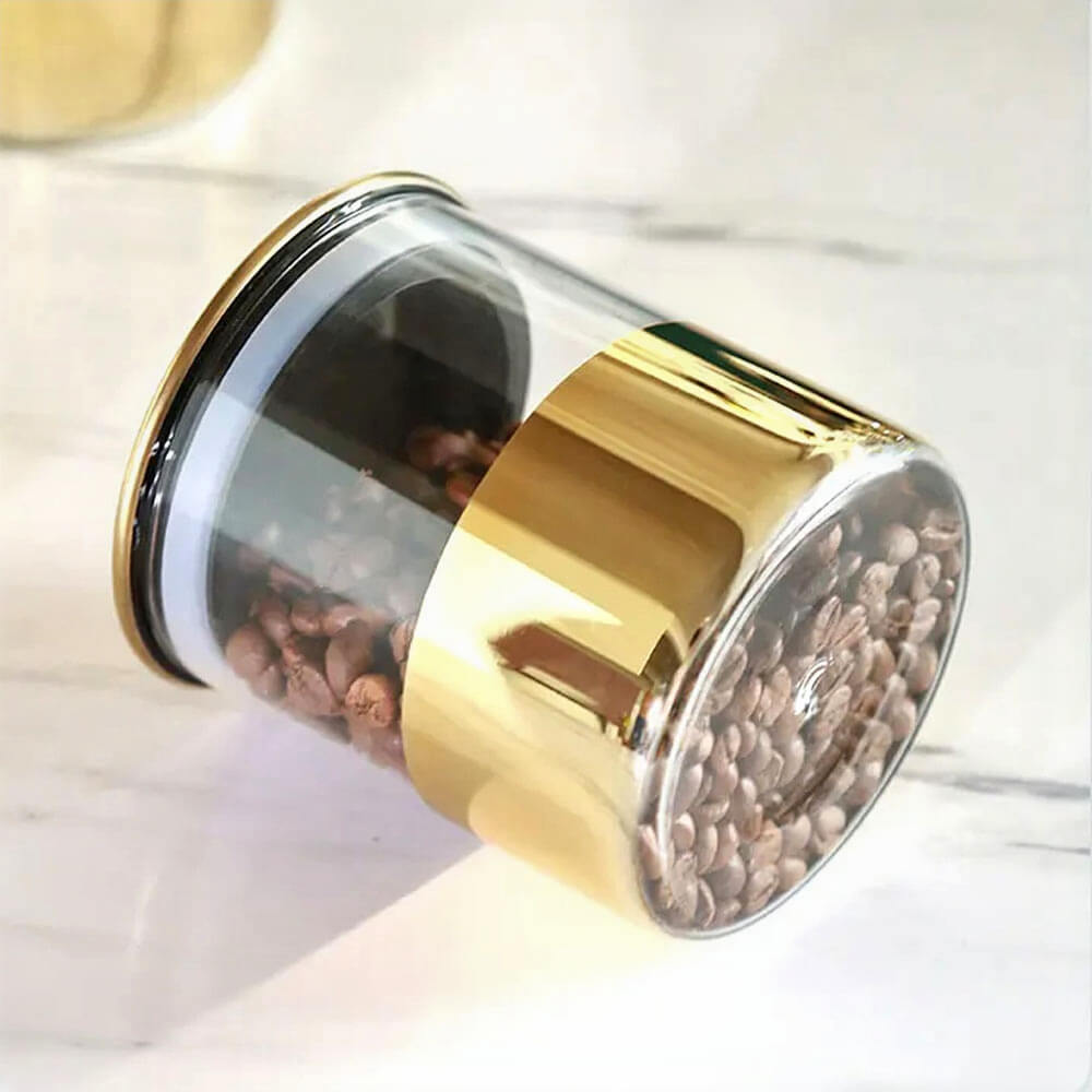 A sleek and modern stackable glass kitchen canister with a gold metallic base and lid is displayed on a marble countertop. The canister is filled with coffee beans, emphasizing its airtight design that keeps contents fresh.