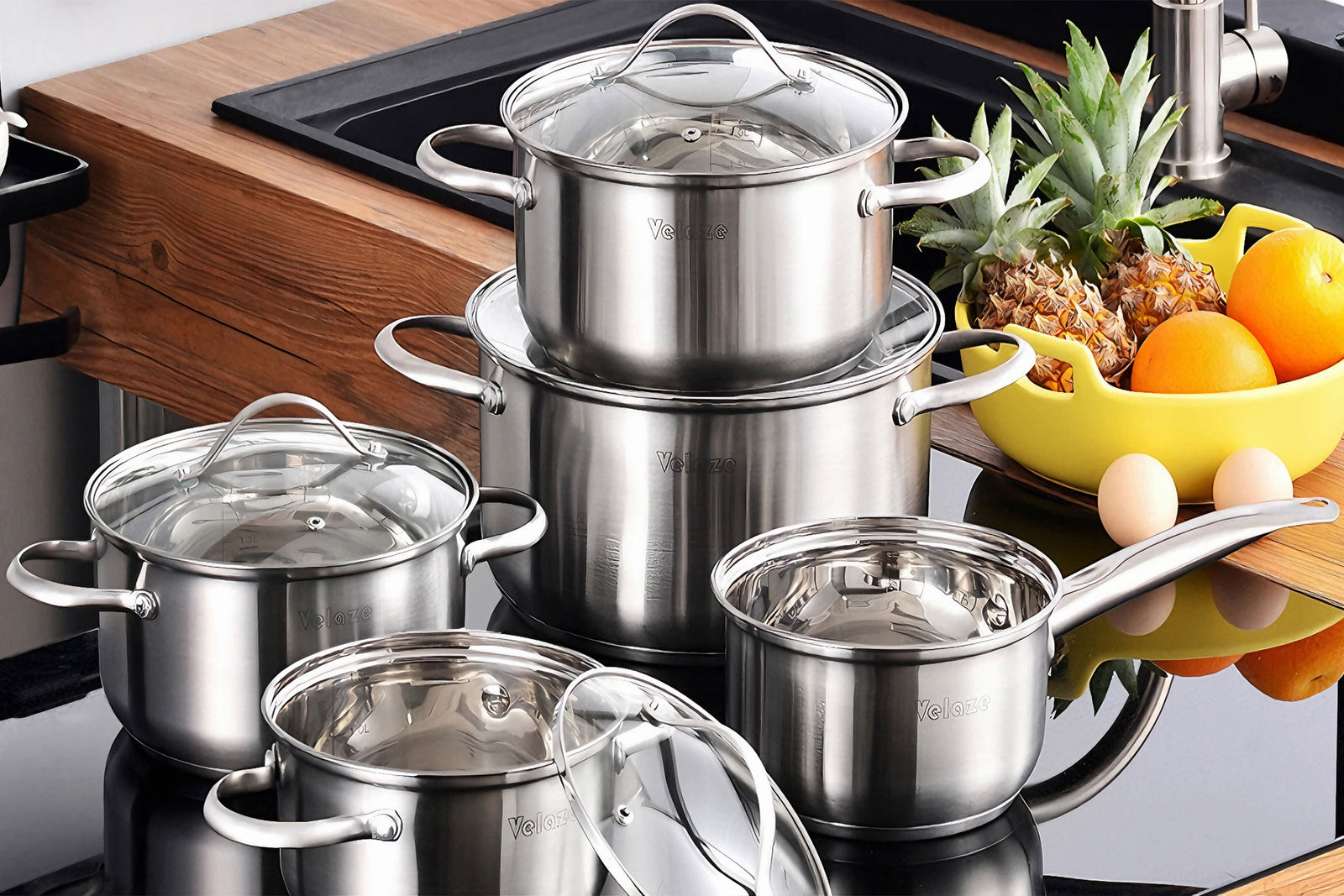 A stylish set of stainless steel cookware with glass lids, featuring various pots and a saucepan, placed on a modern kitchen countertop. A yellow fruit bowl with pineapples, oranges, and eggs sits nearby, enhancing the aesthetic of the cooking space