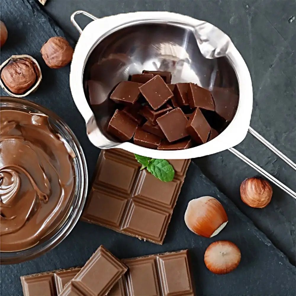Stainless steel double boiler with chocolate pieces inside, surrounded by chocolate bars, melted chocolate, and hazelnuts.