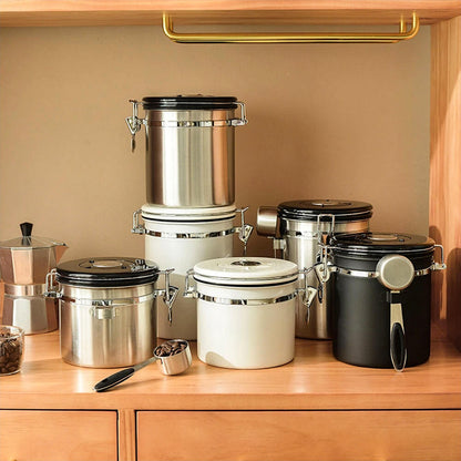 A collection of stainless steel food storage containers with lids in various sizes and colors, featuring airtight seals, secure latches, and attached spoons, neatly arranged on a wooden countertop in a warm kitchen setting.