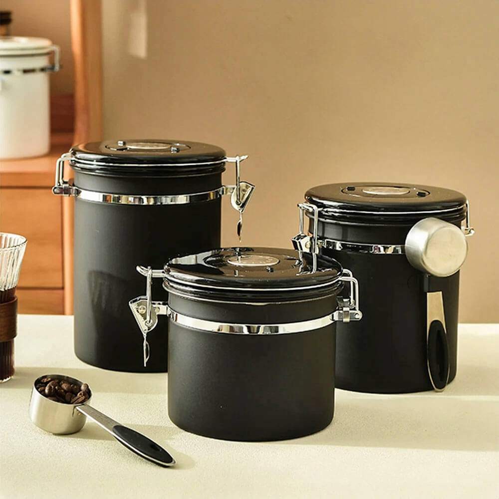 A set of stainless steel food storage containers in matte black, featuring airtight lids, secure latches, built-in date trackers, and attached spoons, displayed on a beige countertop with a coffee scoop filled with beans.