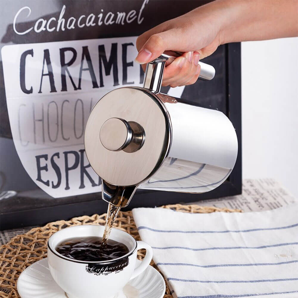 Stainless steel french press coffee maker pouring fresh coffee into a white cup on a woven placemat.