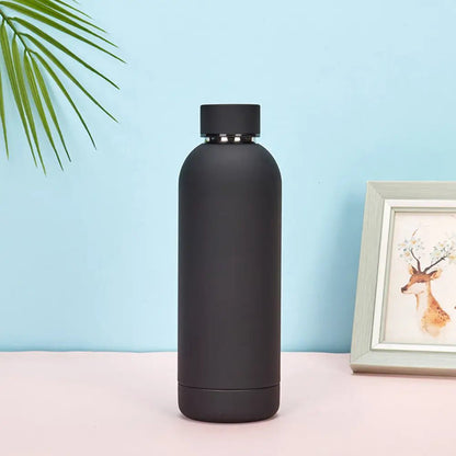 Black water bottle on a pink surface with a light blue background and a framed picture of a deer.