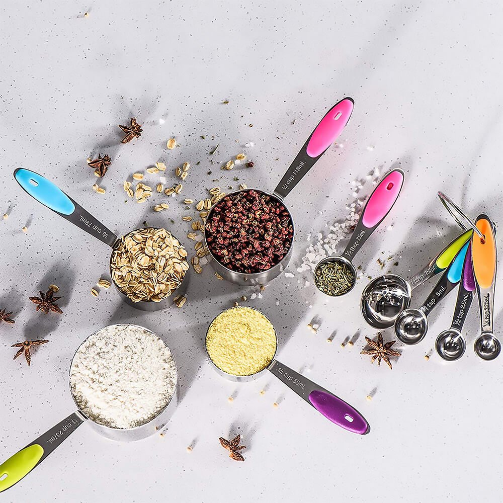 Top view of a stainless steel measuring cup and spoon set filled with ingredients like oats, flour, spices, and herbs. The set features colorful non-slip silicone handles, engraved measurements, and a removable ring holding the spoons together.