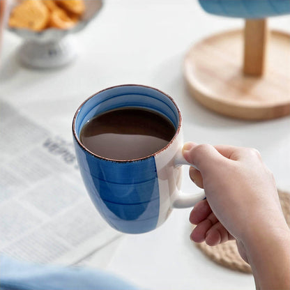 A hand holds a blue stoneware coffee mug filled with a dark beverage, likely coffee. The mug features a smooth finish with subtle stripes and a rustic rim. The background includes a blurred view of a newspaper, a wooden coaster, and some snacks.