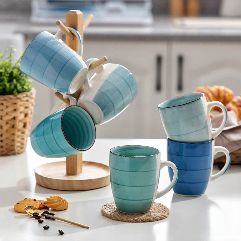 A collection of stoneware coffee mugs in various shades of blue and green is displayed on a white table. Some mugs hang on a wooden mug tree, while others are stacked or placed upright. The mugs feature a striped design with a rustic rim. The background includes kitchen elements, a woven basket with greenery, and a plate of pastries.