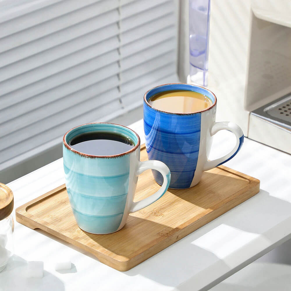 Two stoneware mugs from a stoneware mug set are placed on a wooden tray in a well-lit setting. One mug is light blue with black coffee, and the other is deep blue with a lighter beverage, possibly tea or latte. The mugs feature a striped design and rustic rims. The background includes a coffee machine and soft natural lighting from blinds, creating a cozy and refreshing scene.