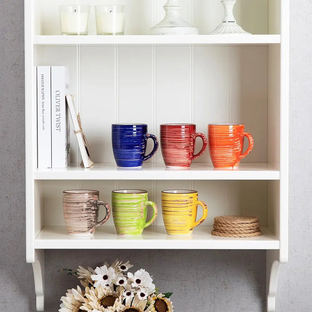A set of six colorful stoneware mugs displayed on a white shelf, featuring red, blue, orange, beige, green, and yellow finishes.