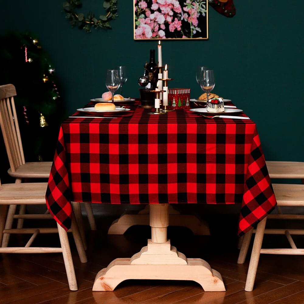 A cozy dining table features a vibrant red and black tartan plaid tablecloth, creating a warm and festive atmosphere. The table is set with wine glasses, white plates, and small dessert servings, accompanied by elegant candles in gold holders. Wooden chairs and a green wall with floral decor in the background complete the inviting setup, ideal for holiday gatherings.